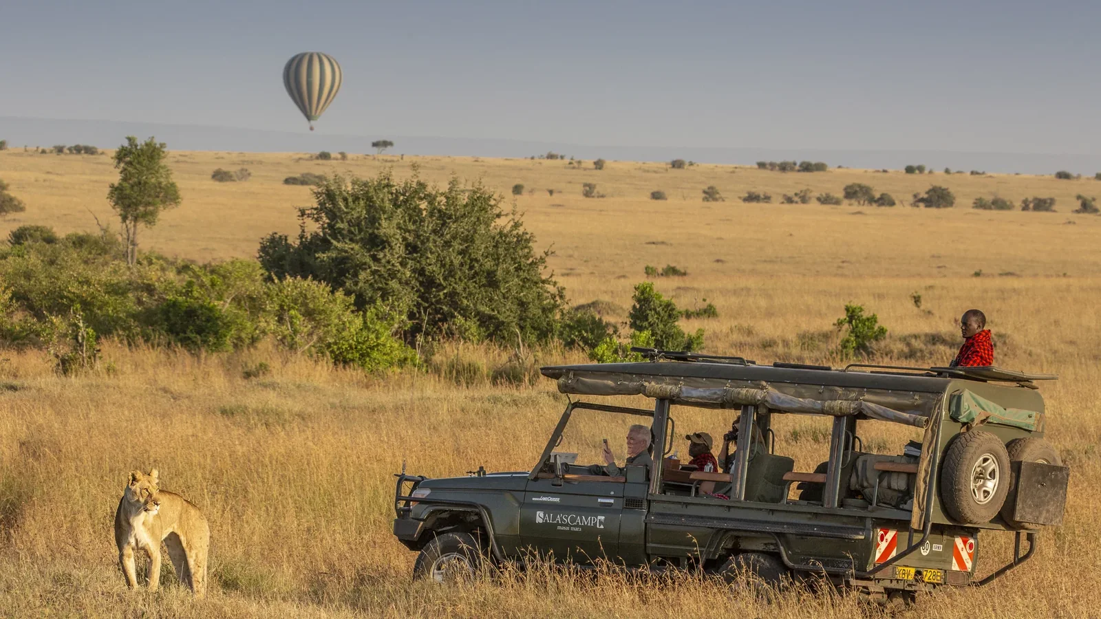 Lioness beside a Sala's Camp safari vehicle with a hot-air balloon above the Maasai Mara