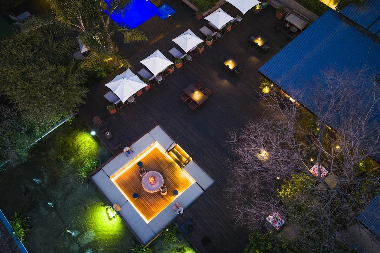 Overhead view of a lit fire pit and sunken seating beside a terrace with white umbrellas and a blue-lit pool.