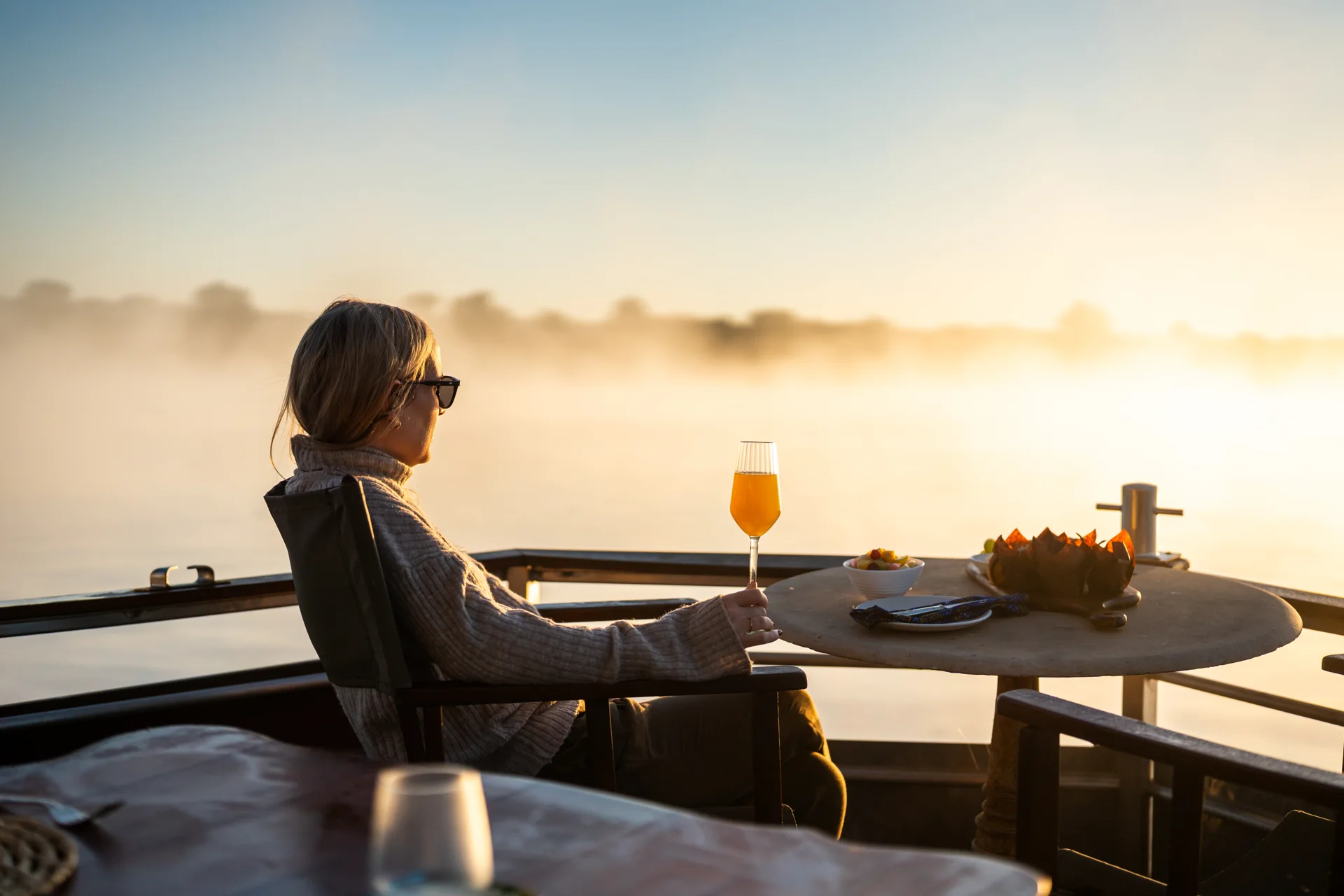 Guest holding a glass beside a breakfast table on a boat overlooking a misty river.
