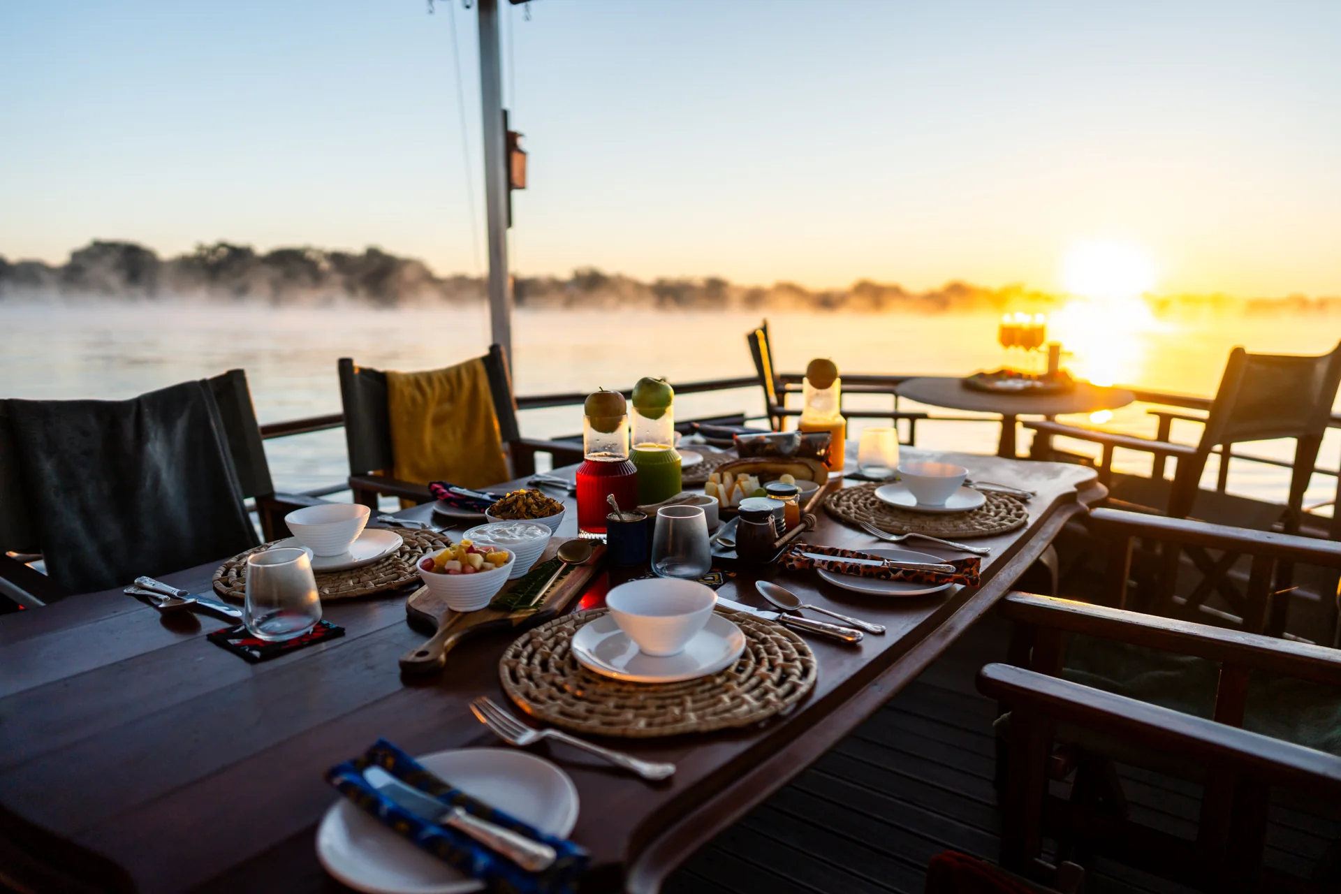 Breakfast table set with fruit, juice and bowls on a boat, with sunlight over the misty river.