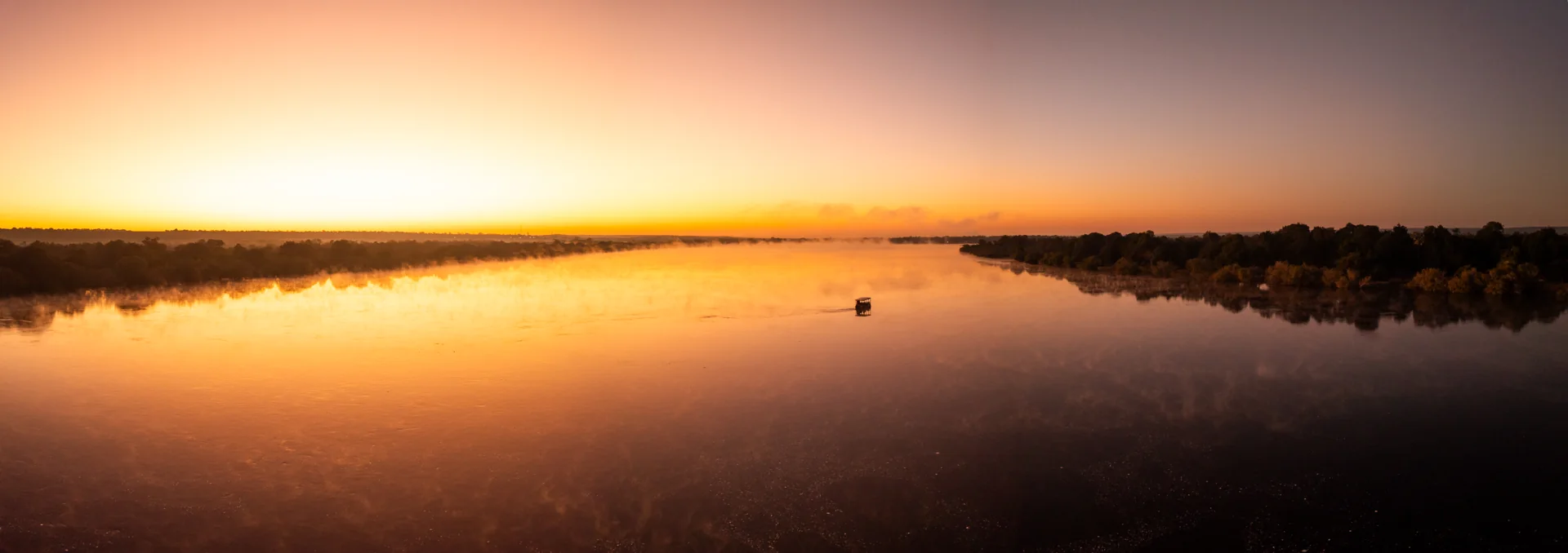 Small boat crossing a broad, misty river beneath a golden sky.